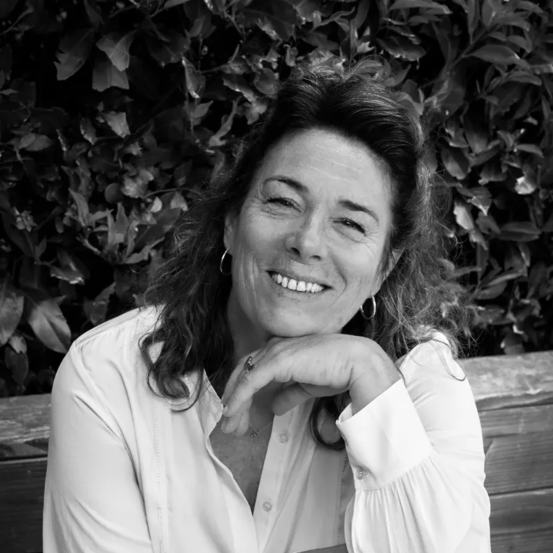 Portrait de Lise Baussan en noir et blanc, sur fond feuillis, avec les cheveux à moitié attachés, un haut blanc et la main gauche sous le menton