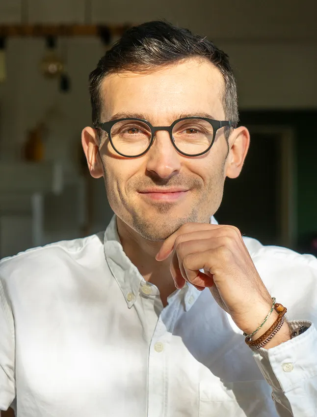 Portrait de Bertrand Deriot, porte une chemise blanche, des lunettes de vue, se tient le menton avec la main gauche et sourit légèrement